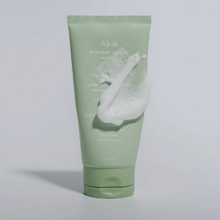 Grön tub med Abib Acne Foam Cleanser Heartleaf, täckt av ett vitt skum som illustrerar den skummande rengöringseffekten. Produkten innehåller hjärtbladsextrakt och salicylsyra för aknebenägen hud.