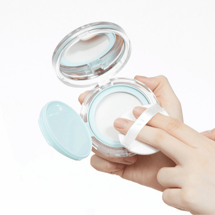 En hand håller en öppen burk med VT PDRN Essence Glow Sun Pact. Puffen är på väg att applicera pudret, som har en vit yta och en blå kant runt burken.