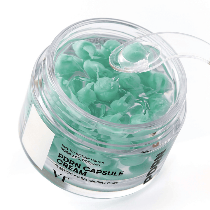 Närbild på en öppen burk med VT PDRN Capsule Cream. Innehållet består av små gröna kapslar i en genomskinlig gel, med en medföljande spatel för applicering. Texten på burken lyder "Elasticity & Balancing Care".