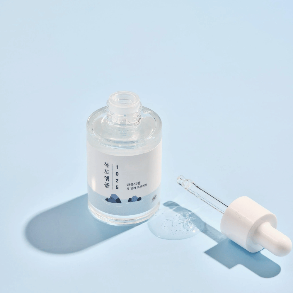 Een open fles van 1025 Dokdo Ampoule met een druppelaar ernaast, tegen een blauwe achtergrond die een zachte schaduw creëert.