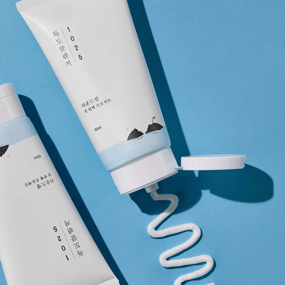 Twee tubes van 1025 Dokdo Cleanser op blauwe achtergrond, waarvan een met open dop en een golvende streng reiniging uitgeperst.