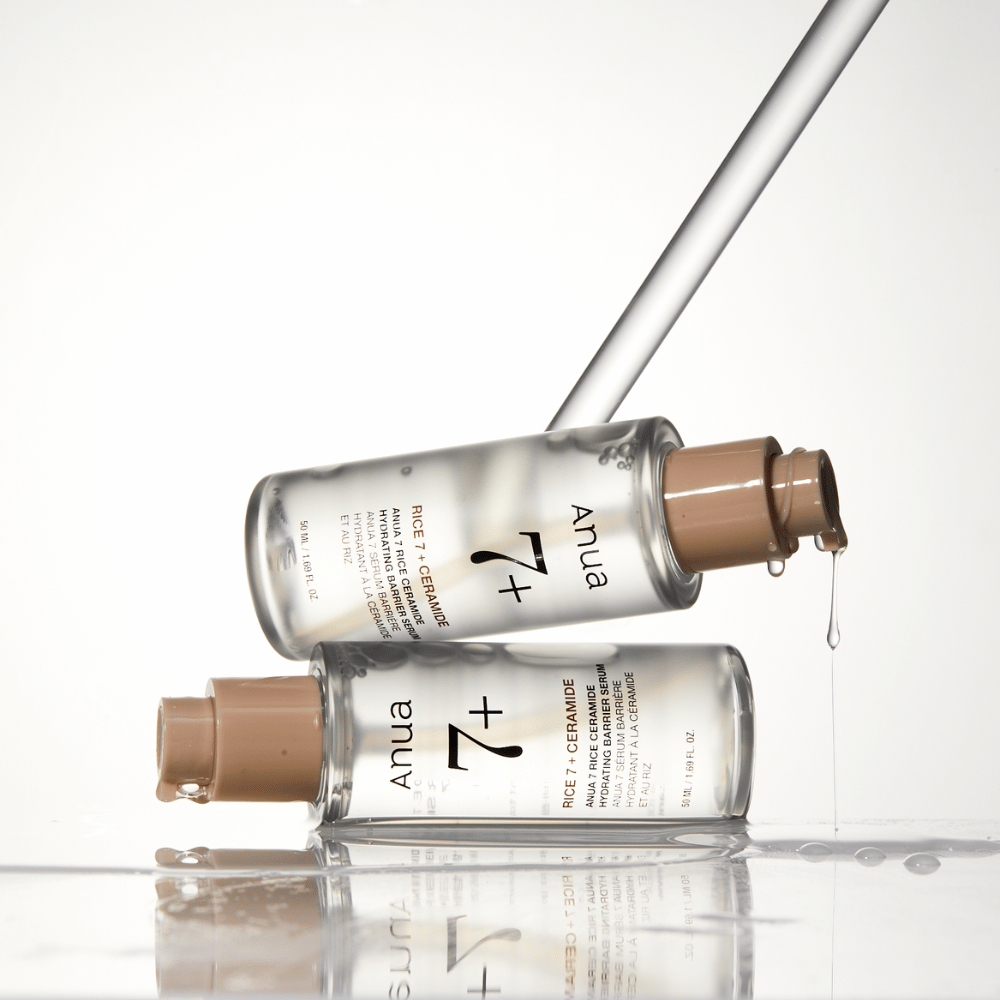 De afbeelding toont twee doorzichtige flessen van "Anua 77 Rice + Ceramide Hydrating Barrier Serum" met druppels die van een pipet lopen. Het serum, dat verrijkt is met rijst en ceramiden, is bedoeld om de huidbarrière te hydrateren en te versterken. De flessen zijn elegant ontworpen, wat het premiumgevoel van het product versterkt.
