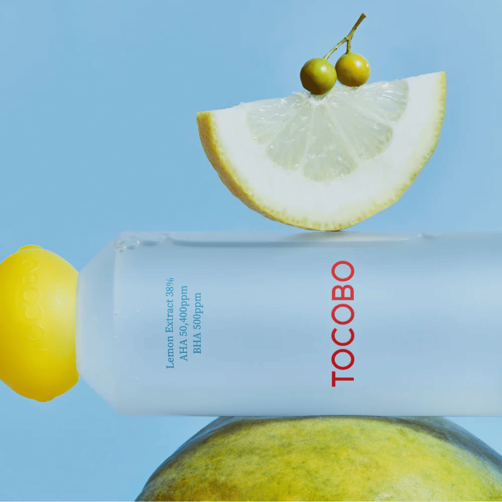 Fles met TOCOBO AHA BHA Lemon Toner, versierd met een dop in de vorm van een citroen. Een schijfje citroen en kleine groene bessen balanceren bovenop de fles tegen een blauwe achtergrond.