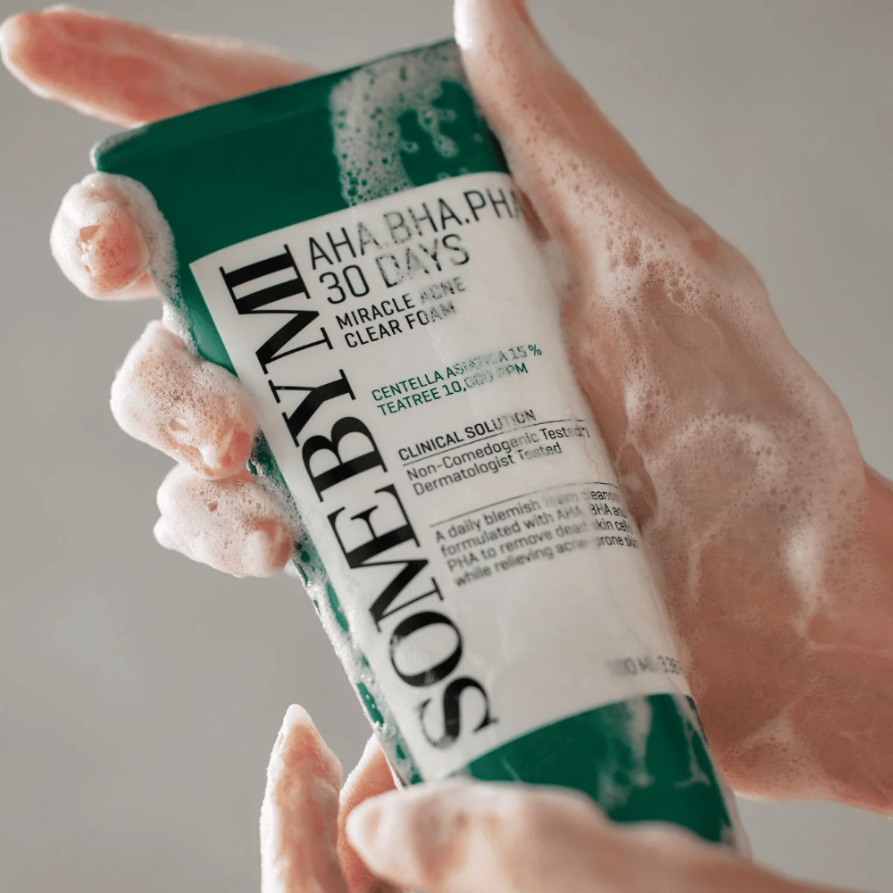 Een persoon houdt een tube van "SOME BY MI AHA, BHA, PHA 30 Days Miracle Acne Clear Foam" met schuimend inhoud over de handen. Het product is bedoeld voor dagelijks gebruik om acne te bestrijden en de poriën te reinigen met zijn actieve ingrediënten zoals centella, tea tree olie en zuren zoals AHA, BHA en PHA.