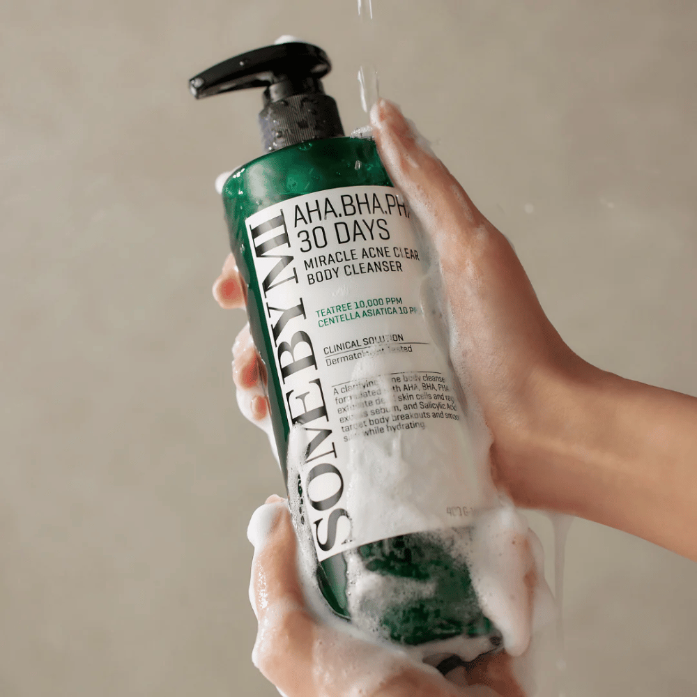 Een persoon houdt een fles SOME BY MI AHA.BHA.PHA 30 Days Miracle Acne Clear Body Cleanser vast. De fles is groen met een wit etiket en is bedekt met schuim van het reinigingsmiddel. De persoon wast zich met het product terwijl er water over de handen stroomt.