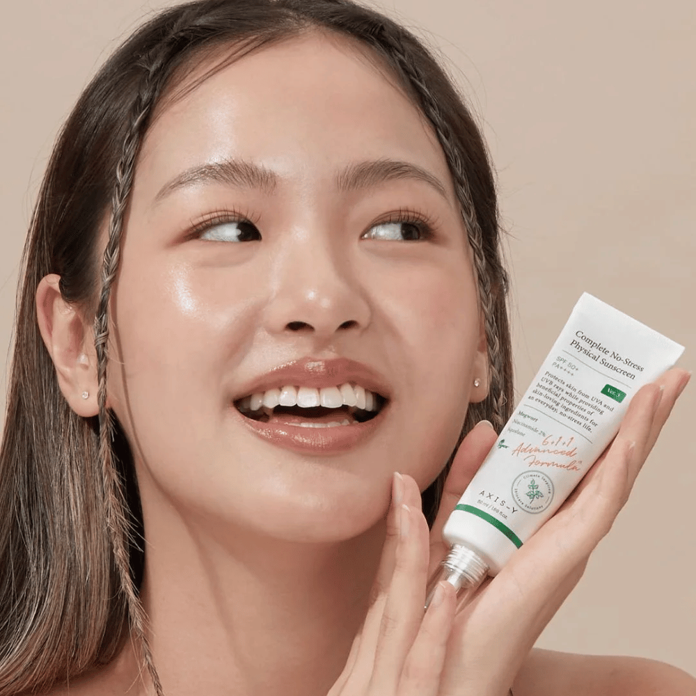 De afbeelding toont een jonge vrouw die vrolijk lacht en een tube vasthoudt met AXIS-Y Complete No-Stress Physical Sunscreen. Ze heeft een stralend en natuurlijk uiterlijk met minimale make-up, wat het product dat ze presenteert benadrukt.