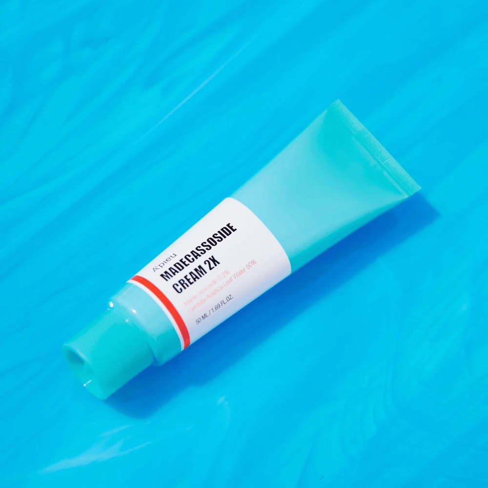 Een tube A'PIEU Madecassoside Cream 2X ligt diagonaal over een blauwe achtergrond met waterachtige rimpels. De tube heeft een witte body met een licht turquoise dop en een rood tot oranje gekleurde etiket waarop de productnaam en het actieve ingrediënt Madecassoside 2% duidelijk staan vermeld. Op het etiket staat ook de hoeveelheid van 50 ml / 1.8 fl oz. De lichte en kleurrijke presentatie geeft een frisse en energieke indruk.