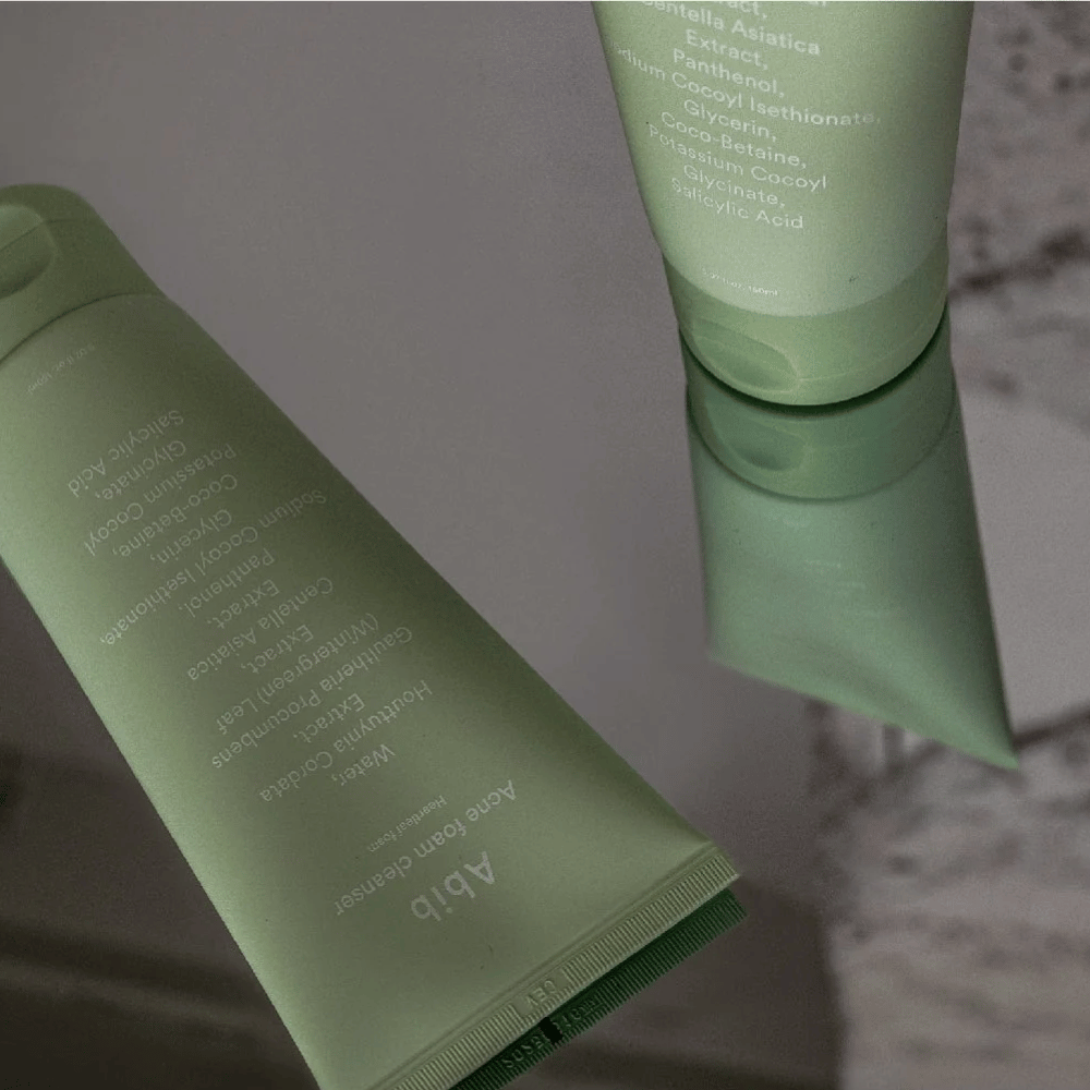 Groene tube van Abib Acne Foam Cleanser Heartleaf liggend op een spiegeltafel met de voorkant van het product en tekst gespiegeld. Ingrediënten zoals salicylzuur, centella en hartbladoestrakt zijn duidelijk zichtbaar op de verpakking.