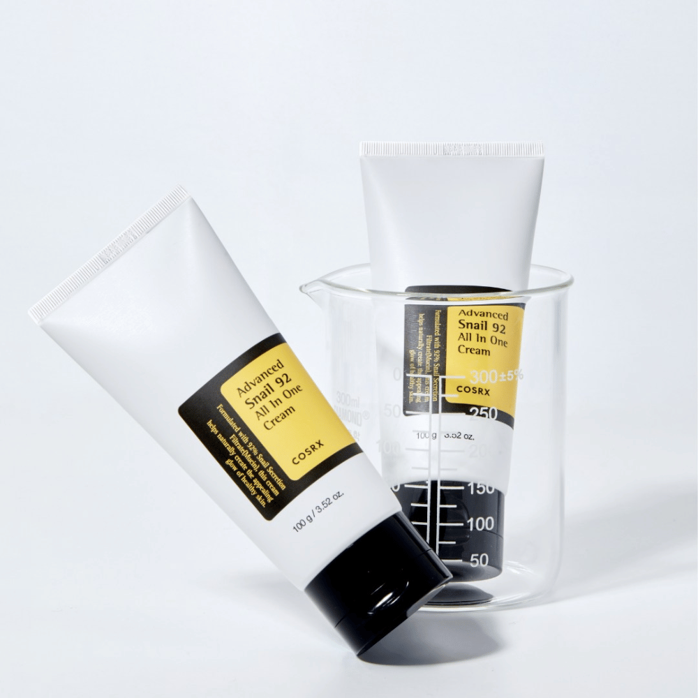 Een tube van COSRX Advanced Snail 92 All in One Cream liggend naast een omgekeerde transparante maatcilinder op een witte achtergrond.