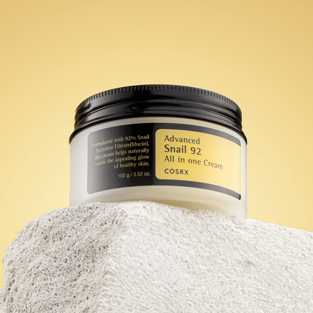 Een pot van COSRX Advanced Snail 92 All in one Cream geplaatst op een rotsachtig oppervlak met een gele achtergrond.