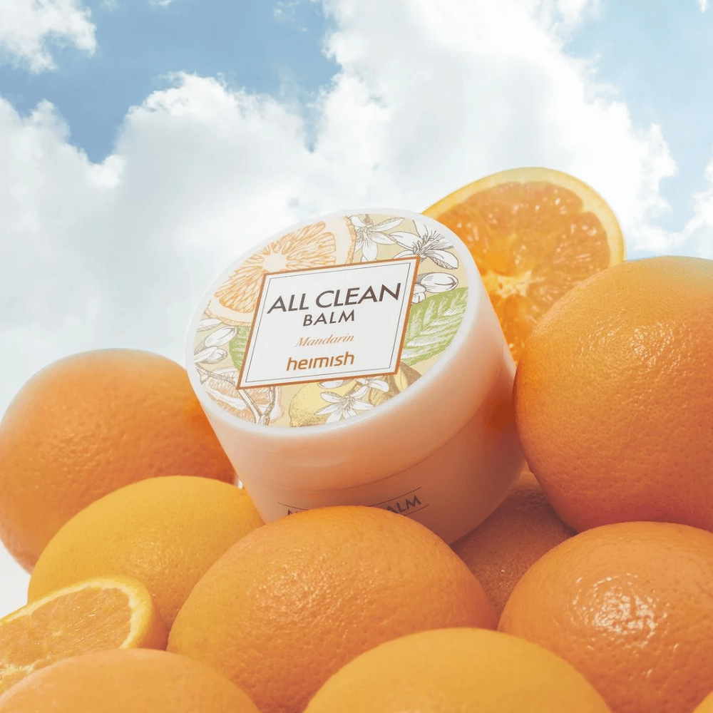 Pot van Heimish All Clean Balm Mandarin geplaatst op een stapel sinaasappels, met een blauwe hemel op de achtergrond.