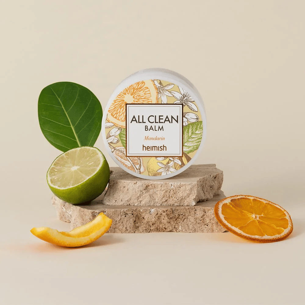 Pot van Heimish All Clean Balm Mandarin geplaatst op stenen, omringd door mandarijnenschillen en citrusbladeren, tegen een lichte achtergrond.
