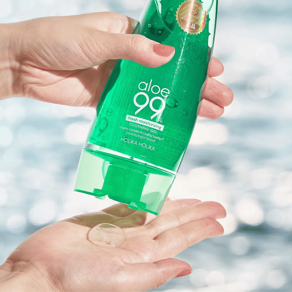 Een hand houdt een groene tube met Holika Holika Aloe 99% Soothing Gel, terwijl een andere hand een druppel gel opvangt die wordt gedoseerd. De achtergrond is wazig en toont een waterig, glinsterend patroon.