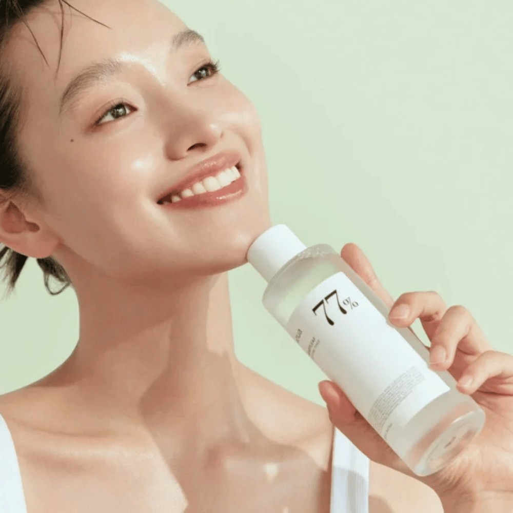 Een vrouw kalmeert haar gevoelige huid met een fles Heartleaf 77% Soothing Toner van Anua, met extract van kameleontblad.