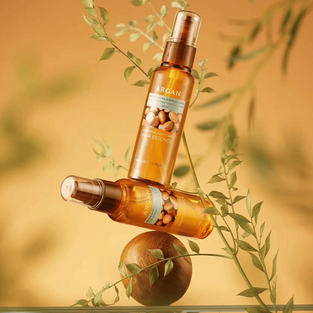 Twee flessen Nature Republic Argan Essential Moisture Hair Oil geplaatst tussen groene bladeren – zijdezachte glans voor droog haar.