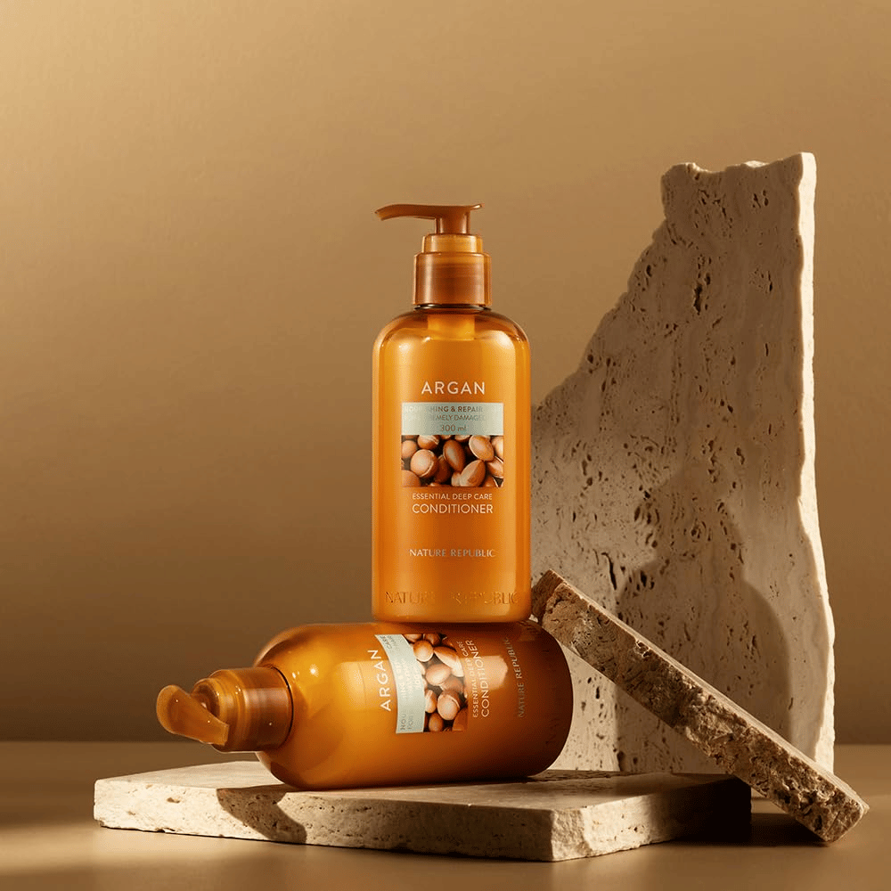 Twee flessen van Nature Republic Argan Essential Moisture Repairing Conditioner op steenoppervlak in warme tint.