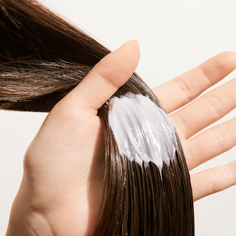 Hand brengt Nature Republic Argan Essential Moisture Repairing Hair Mask aan in nat haar – hydraterend masker dat het haar versterkt en verzacht.