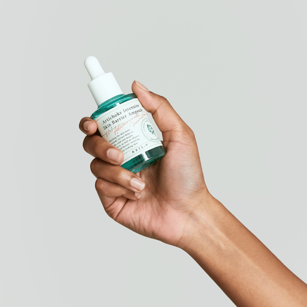 Een hand houdt een fles huidverzorgingsserum met het etiket "Artichoke Intensive Skin Barrier Ampoule.