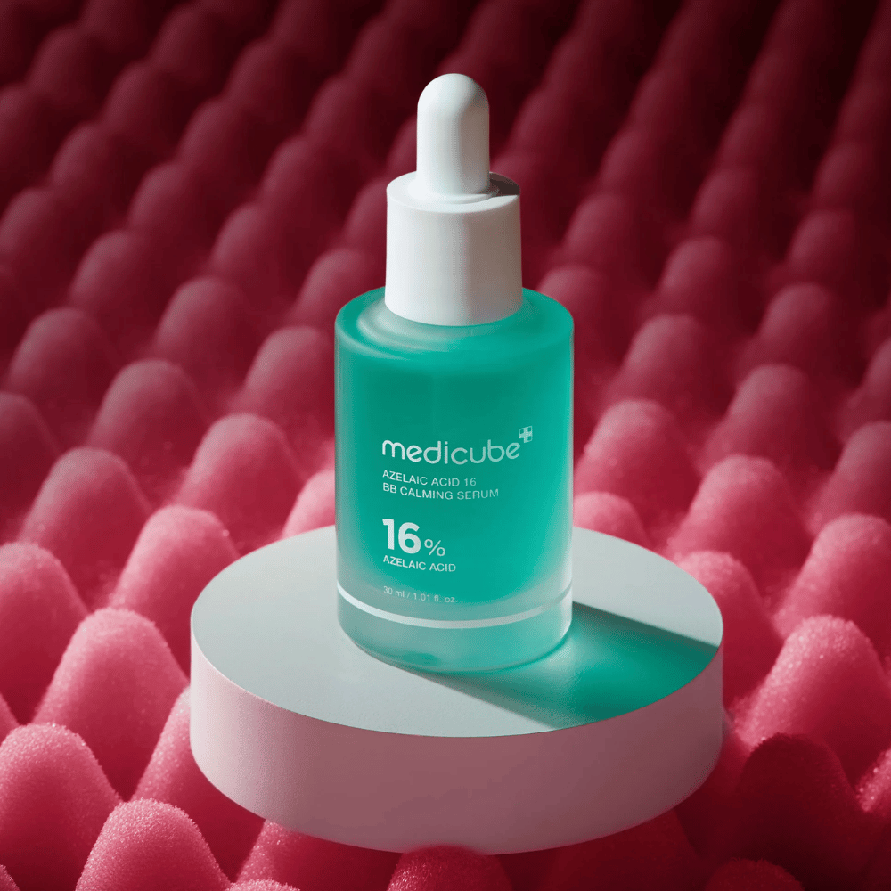 Productafbeelding van Medicube Azelaic Acid 16 BB Calming Serum geplaatst op een wit podiumoppervlak met een roze gestructureerde achtergrond die het kalmerende en huidverbeterende profiel van het serum benadrukt.