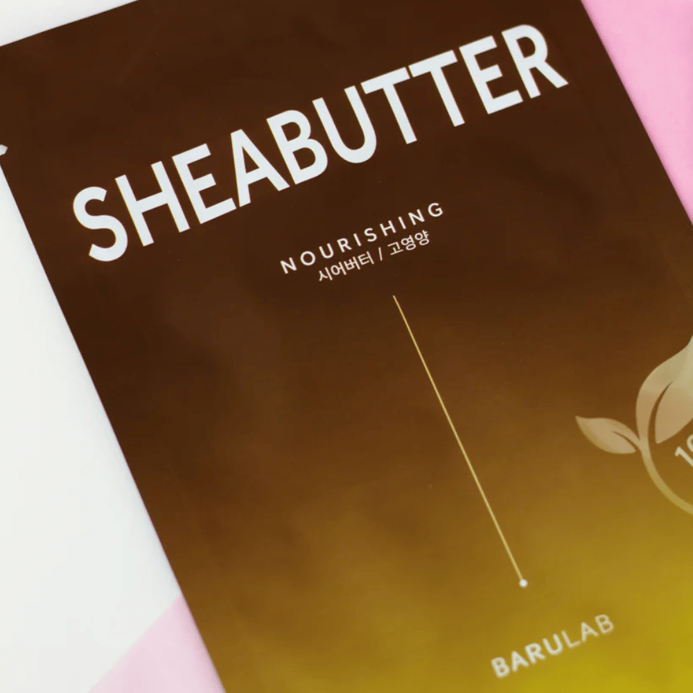 Een close-up van een verpakking van BARULAB "SHEABUTTER Nourishing" masker. Het product is gemarkeerd als "100% Vegan Product" en heeft een kleurverloop van donkerbruin naar lichtbruin.