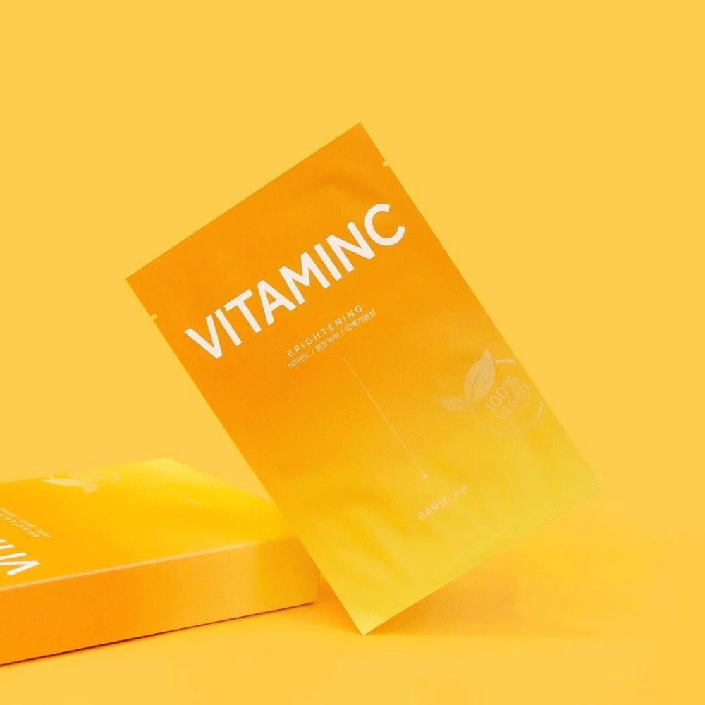 Een stapel verpakkingen van BARULAB "VITAMINC Brightening" maskers, allemaal gemarkeerd als "100% Vegan Product". De verpakkingen hebben een kleurtint in verschillende tinten oranje en geel, geplaatst tegen een uniforme gele achtergrond.