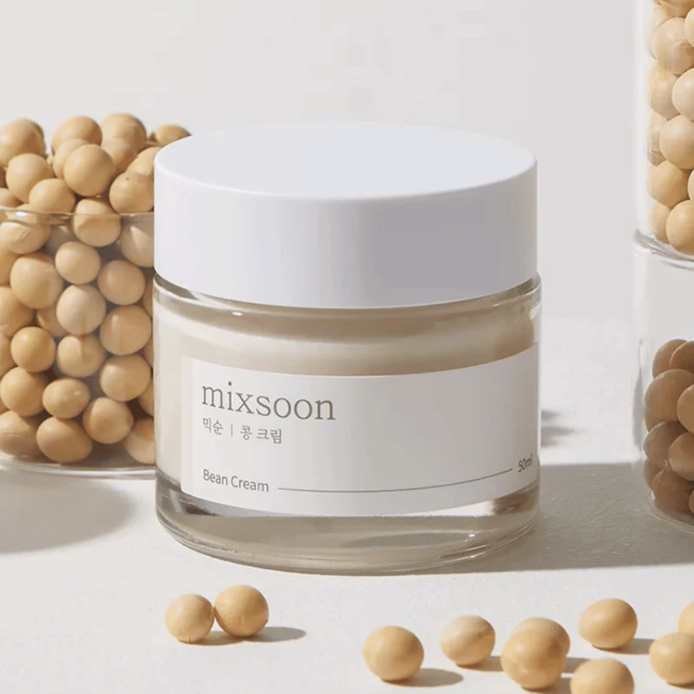 Een doorschijnende pot met wit etiket en deksel, geëtiketteerd "mixsoon 팥 크림" en "Bean Cream" met een aangegeven volume van "50ml". De pot bevat een lichtgele crème en is geplaatst op een lichte ondergrond. Naast de pot staat een glazen houder vol met lichtbruine bonen die over de ondergrond zijn uitgestrooid, wat suggereert dat de crème bonenextract bevat. De achtergrond en belichting geven een warme en natuurlijke sfeer.