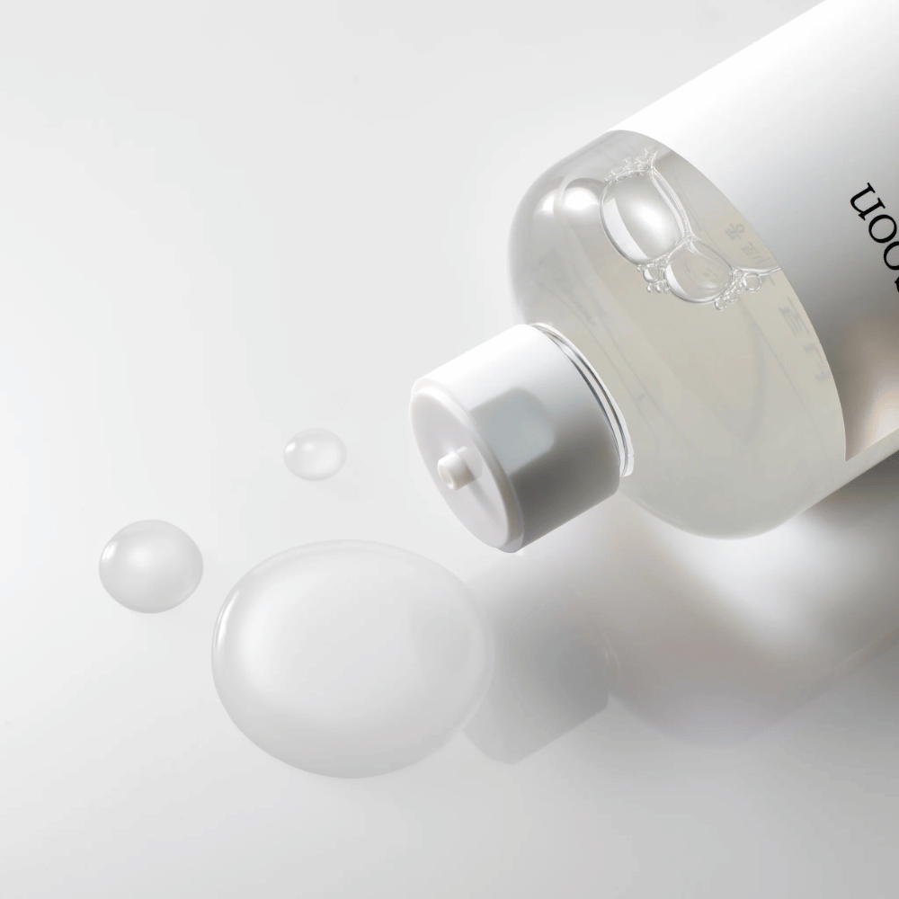 Een doorzichtige fles ligt op zijn kant op een glanzend oppervlak met een lichte toner die langzaam uitstroomt en druppels en een grotere plas op het oppervlak vormt. De fles heeft een wit etiket en een wit pompmechanisme. De verlichting is zacht en creëert een schone en rustgevende sfeer, terwijl de vloeibare consistentie en helderheid van de toner worden benadrukt door de manier waarop deze zich over het oppervlak verspreidt.