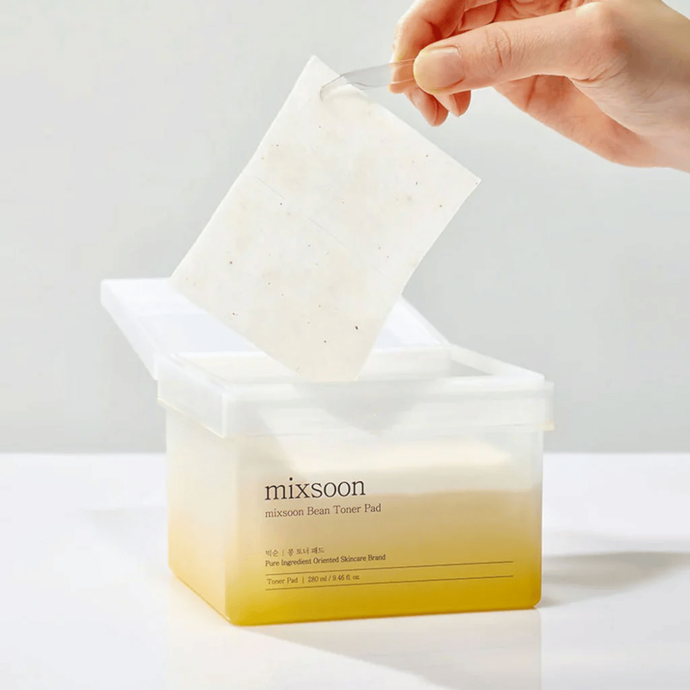 Een hand houdt een pincet vast en tilt een Mixsoon Bean Toner Pad uit een vierkante, transparante container met een gelige tint. De container bevat 70 toner pads en heeft tekst die het product beschrijft.