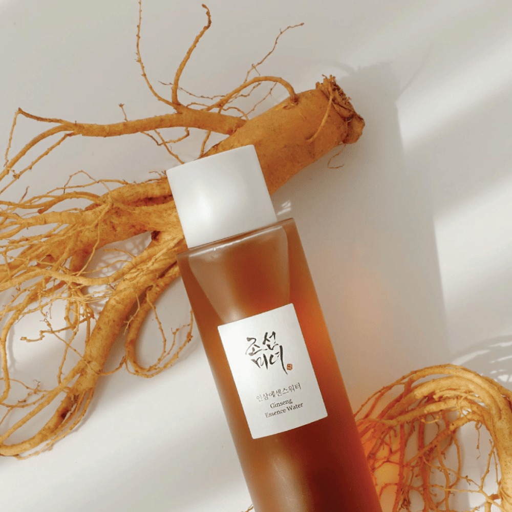 Een bruine fles Beauty of Joseon Ginseng Essence Water met witte dop ligt naast een verse ginsengwortel op witte achtergrond.