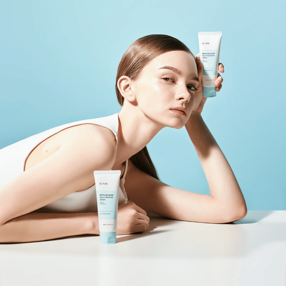Lifestyle-afbeelding waarop een model poseert met iUNIK Beta-Glucan Daily Moisture Cream tegen een lichtblauwe achtergrond. De afbeelding straalt een schoon, fris en hydraterend gevoel uit terwijl het product duidelijk zichtbaar is.