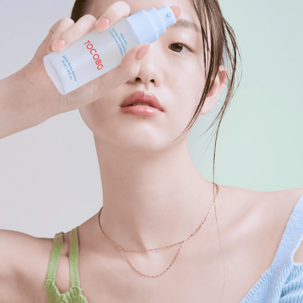 Jonge vrouw met zachte make-up en natuurlijke huidtint houdt een TOCOBO Bifida Biome Essence-fles voor haar gezicht. Ze draagt een lichtblauwe trui met groene schouderbanden en een dunne gouden ketting tegen een lichte achtergrond.