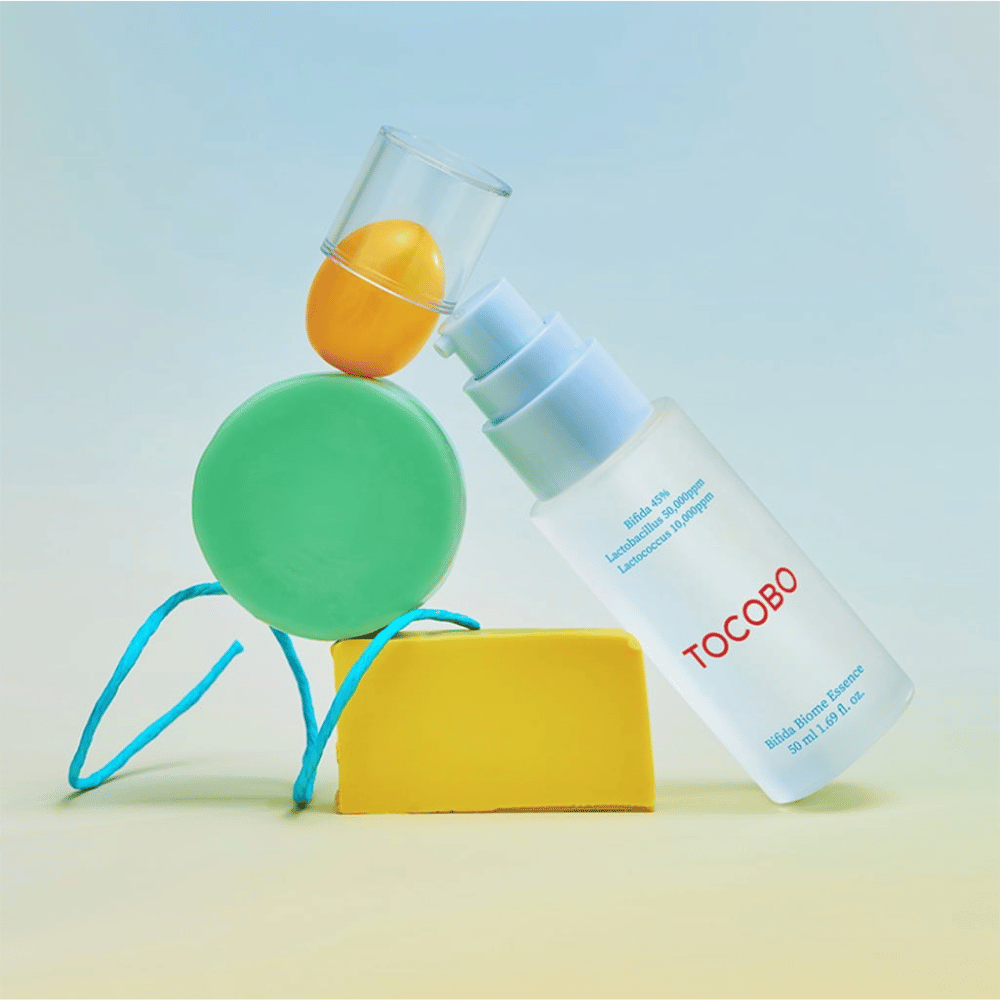 TOCOBO Bifida Biome Essence in matglazen pompflacon met rood logo, geplaatst tegen een kleurrijke achtergrond met groene, gele en oranje geometrische vormen. Tekst op de fles geeft aan Bifida 5%, Lactobacillus 3.000 ppm en Lactococcus 1.000 ppm.