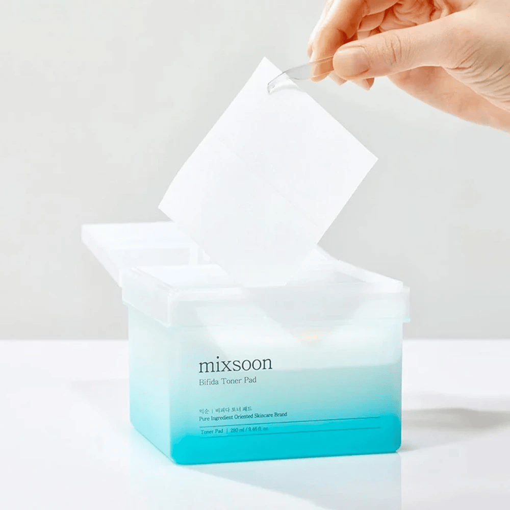 Een transparante container met een blauwe en witte gradient die Bifida toner pads bevat. Een hand neemt een toner pad uit de container, die is gemarkeerd met "mixsoon Bifida Toner Pad" en bevat 280 ml (9,46 fl. oz.) toner pads bedoeld voor huidverzorging.