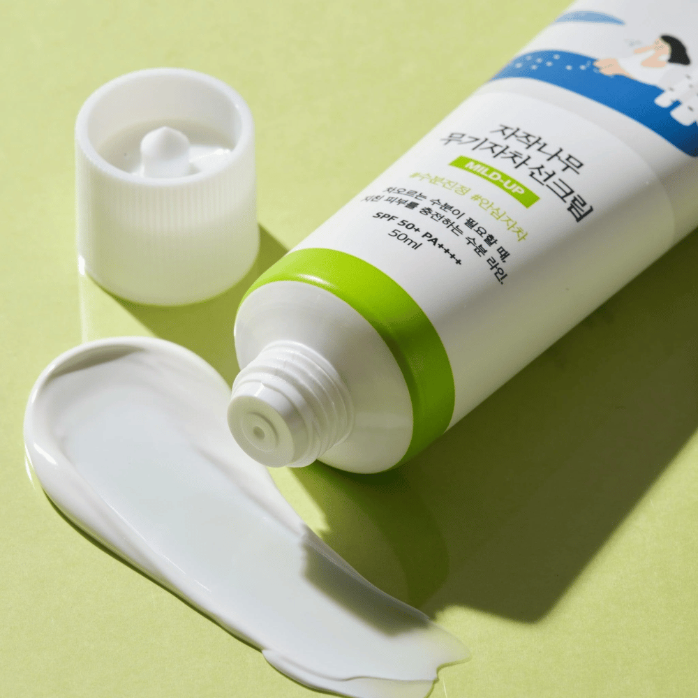 Close-up van Birch Juice Moisturizing Mild Up Sunscreen met losgeschroefde dop, witte zonnebrandcrème uitgesmeerd op lichtgroene achtergrond, SPF 50+ PA++++, 50 ml.