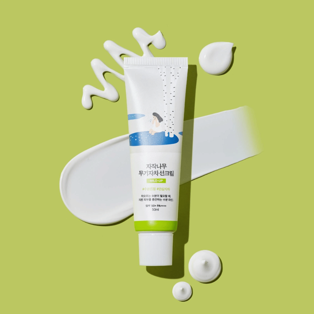Birch Juice Moisturizing Mild Up Sunscreen – witte tube met groene basis op groene achtergrond, omgeven door uitgesmeerde zonnebrandcrème en druppels, SPF 50+ PA++++, 50 ml.