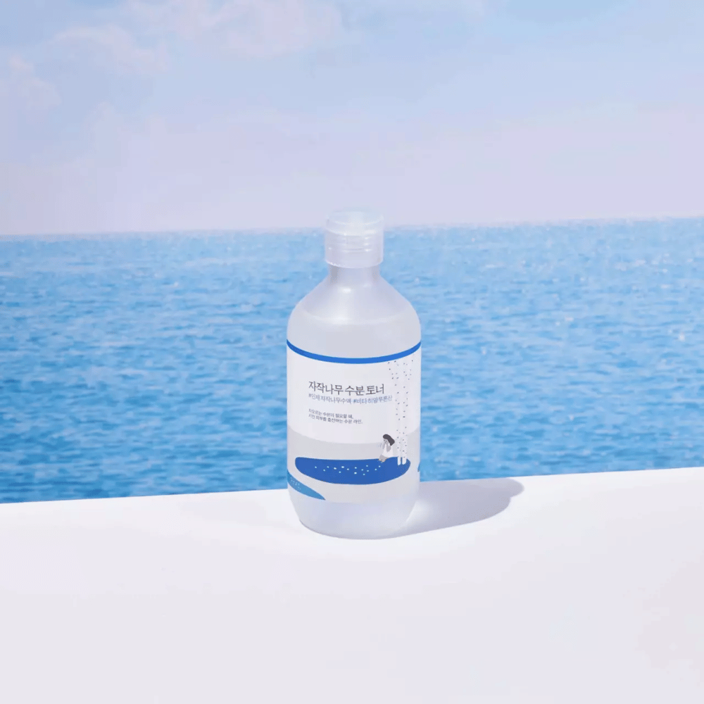 Fles van ROUND LAB Birch Juice Moisturizing Toner staat op een wit oppervlak met zee en blauwe lucht op de achtergrond