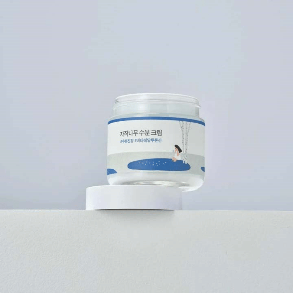 Round Lab Birch Juice Moisturizing Cream in transparante pot met wit etiket en blauwe illustratie van vrouw bij berk, staand op witte ondergrond tegen lichte achtergrond.