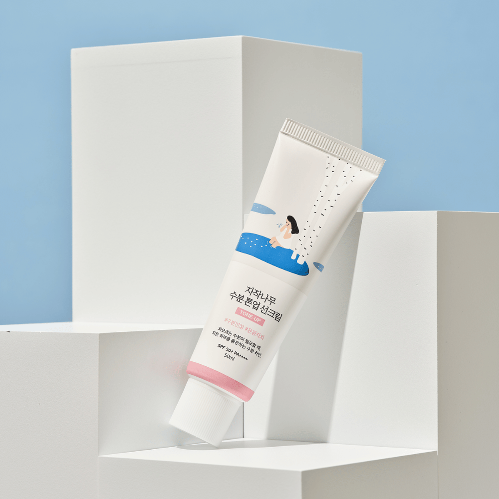 Een tube met Birch Juice Moisturizing Tone Up Sunscreen geplaatst op witte kubussen tegen een lichtblauwe achtergrond. De tube is wit met een illustratie van een vrouw bij een berk en water. De Koreaanse tekst geeft aan dat het product SPF 50+ PA++++ heeft en 50 ml bevat.