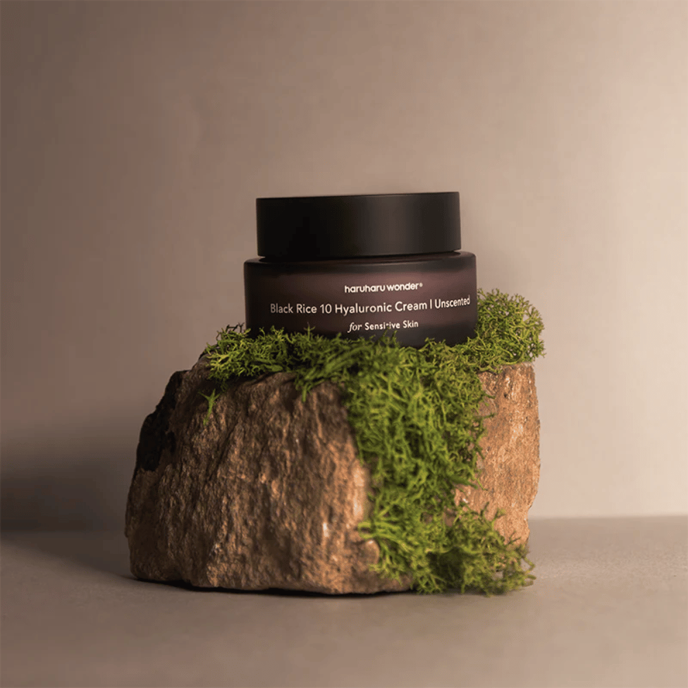 De afbeelding toont een pot huidcrème met het label "haruharu wonder Black Rice 10 Hyaluronic Cream Unscented for Sensitive Skin". De pot is zwart en staat op een grote steen bedekt met lichtgroen mos, wat een natuurlijke en aardse uitstraling creëert.