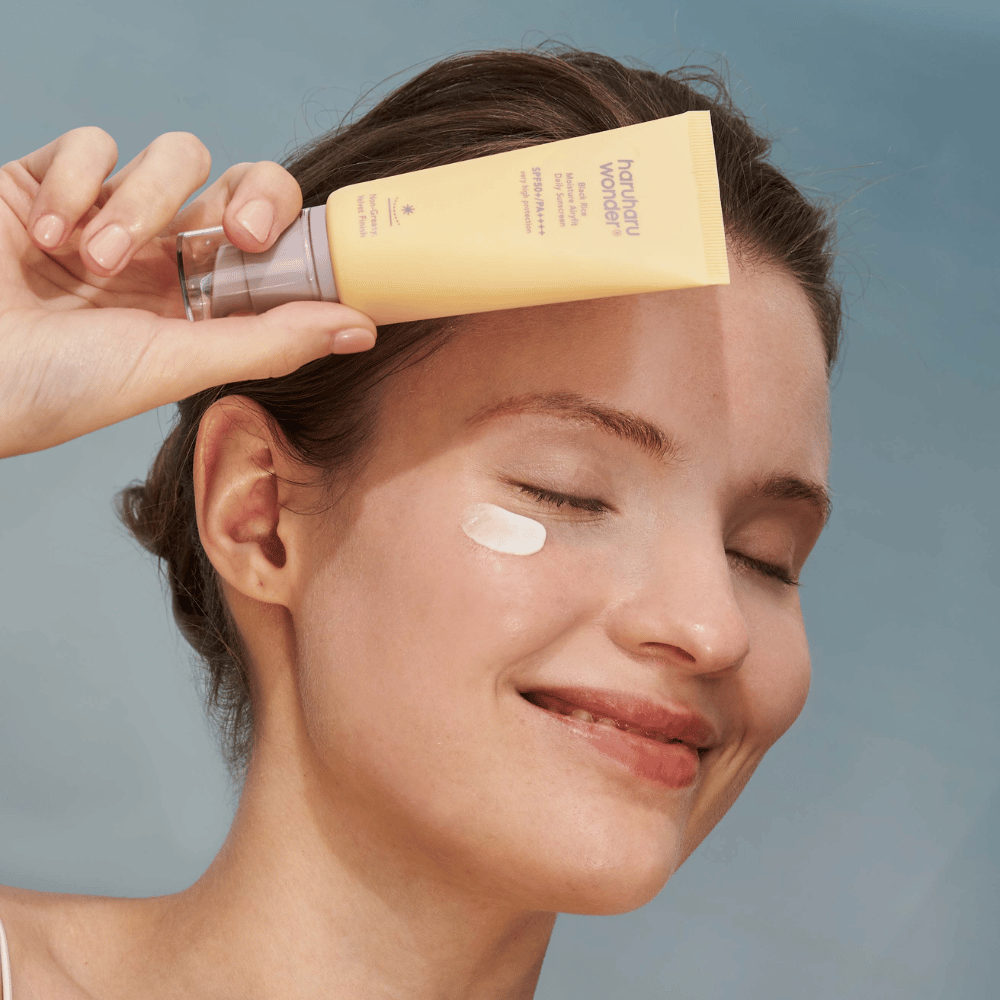 Een glimlachende vrouw brengt Black Rice Moisture Airyfit Daily Sunscreen SPF 50+ PA++++ van Haruharu Wonder aan op haar gezicht. Ze houdt een gele tube met zonnebrandcrème vast en heeft een klodder van het product op haar wang. De achtergrond is lichtblauw.
