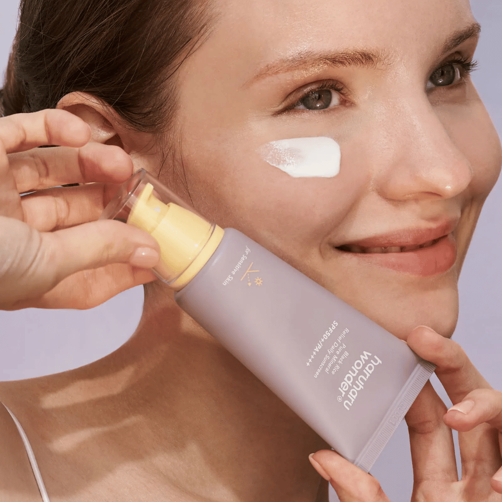 Een glimlachende vrouw brengt Haruharu Wonder Black Rice Pure Mineral Relief Daily Sunscreen SPF 50+ PA++++ aan op haar gezicht. Ze houdt een paarse tube zonnebrandcrème vast en heeft een druppel van het product op haar wang. De achtergrond is lichtpaars.