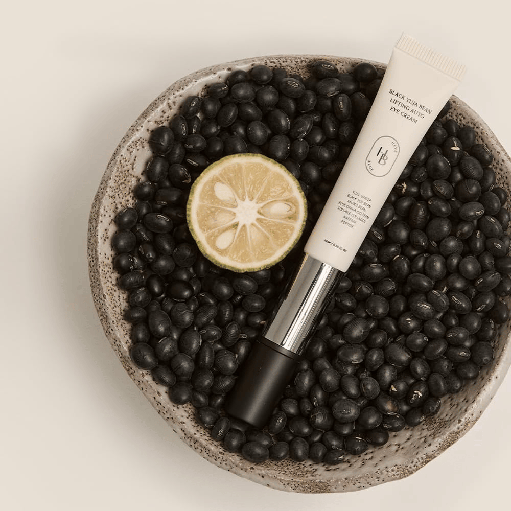 Black Yuja Bean Lifting Auto Eye Cream liggend in een schaal gevuld met zwarte bonen en een gehalveerde citrusvrucht – een visuele representatie van de natuurlijke ingrediënten van het product.