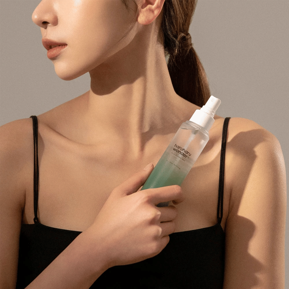 Afbeelding van HaruHaru Wonder Black Bamboo Mist vastgehouden door een vrouw. Het product zit in een transparante spuitfles met een groene tint en een witte pomp. De vrouw heeft een ontspannen uitstraling, draagt een zwarte top en richt zich op het gebruik van de mist om de huid te hydrateren en glans te geven. De fles maakt deel uit van een huidverzorgingsroutine.