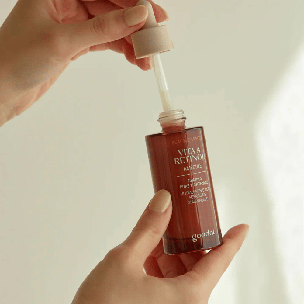 Een hand houdt een fles met Goodal Black Carrot Vita-A Retinol Firming Ampoule vast. Het etiket van de fles toont de naam en eigenschappen van het product, inclusief "Firming Pore Tightening" en ingrediënten zoals 10% hyaluronzuur, adenosine en niacinamide. De pipet is beige van kleur.