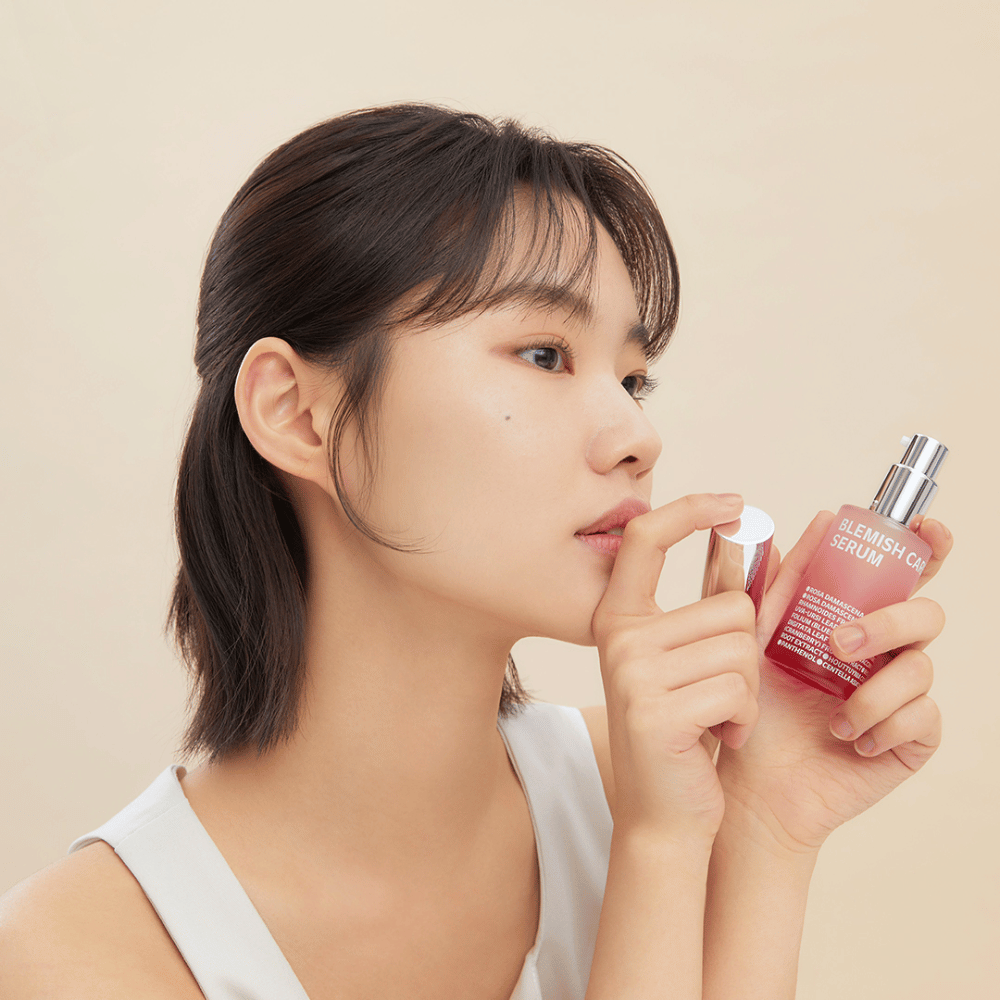 Een vrouw met donker haar houdt een fles Blemish Care Serum in de ene hand en neemt de dop eraf met de andere. Ze heeft een neutrale gezichtsuitdrukking en de achtergrond is licht beige, wat een zachte en minimalistische indruk geeft.