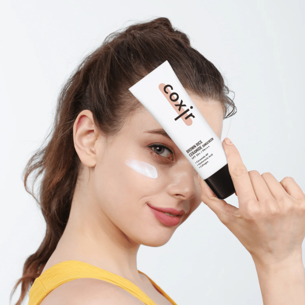 Vrouw met zonnebrandcrème aangebracht op haar wang houdt een tube Coxir Brown Rice Ceramide Sunscreen, SPF 50+ PA+++, tegen haar voorhoofd en glimlacht zachtjes.