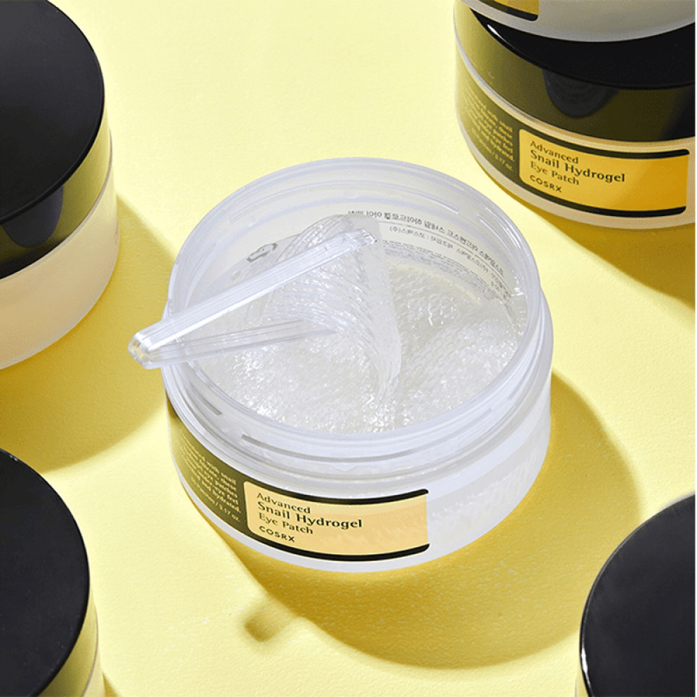 Een open pot COSRX Advanced Snail Hydrogel Eye Patch met doorzichtige oogpatches en een kleine spatel, omringd door meer gesloten potten op een gele achtergrond.