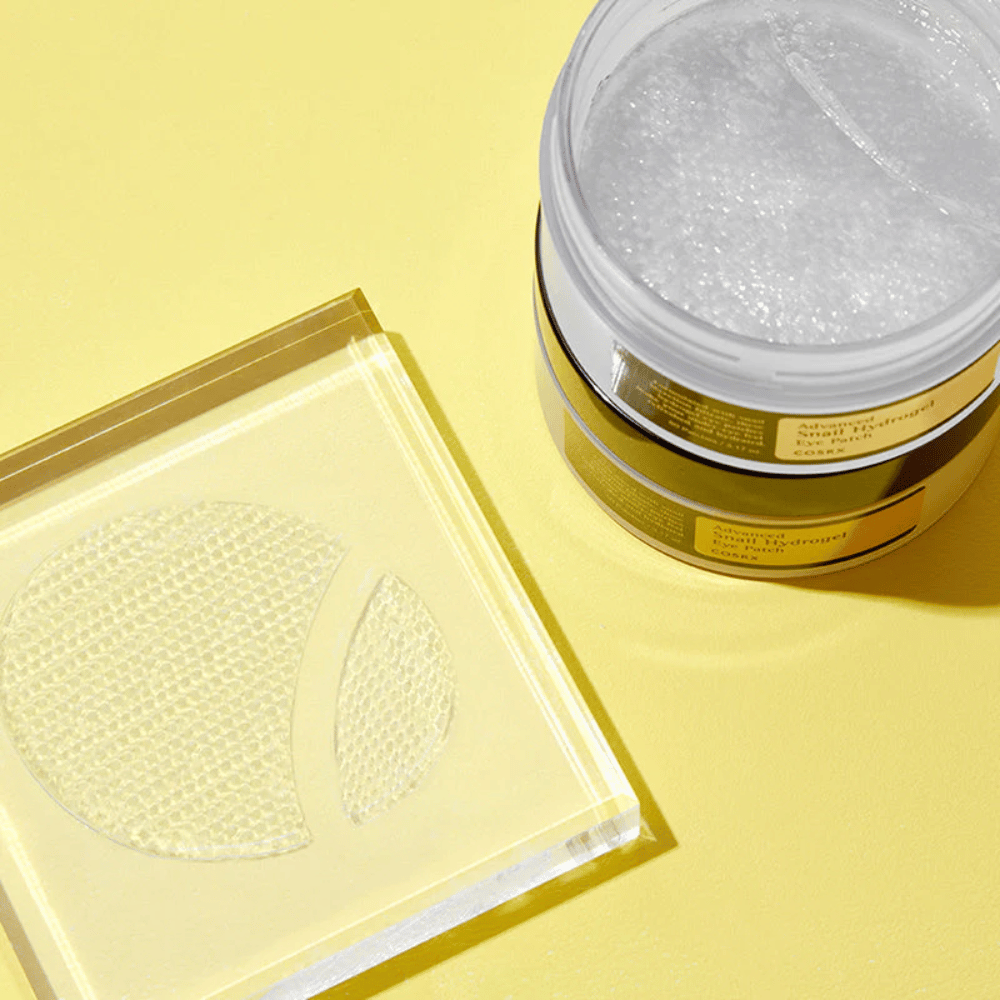 Een open pot COSRX Advanced Snail Hydrogel Eye Patch staat op een gele achtergrond naast een doorzichtig onderzettertje waar twee oogpatches liggen.
