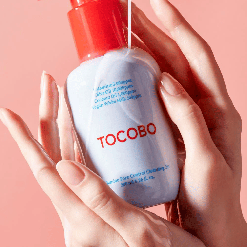 TOCOBO Calamine Pore Control Cleansing Oil wordt in handen gehouden tegen een roze achtergrond, de rode pomp van de fles en de ingrediëntenlijst zijn duidelijk zichtbaar.