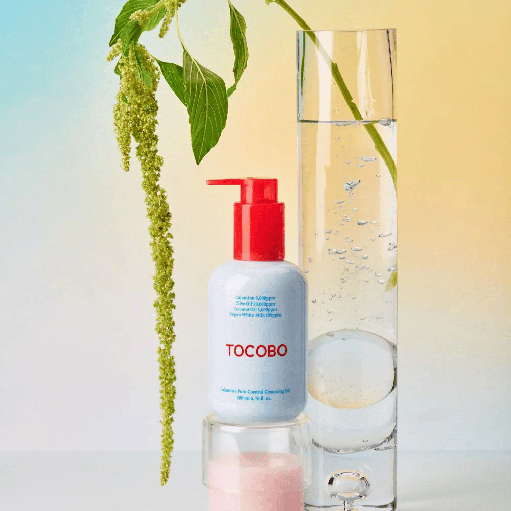 TOCOBO Calamine Pore Control Cleansing Oil met rode pomp staat naast een hoog glazen buis gevuld met water en een groene plant, tegen een lichte pastelkleurige achtergrond.